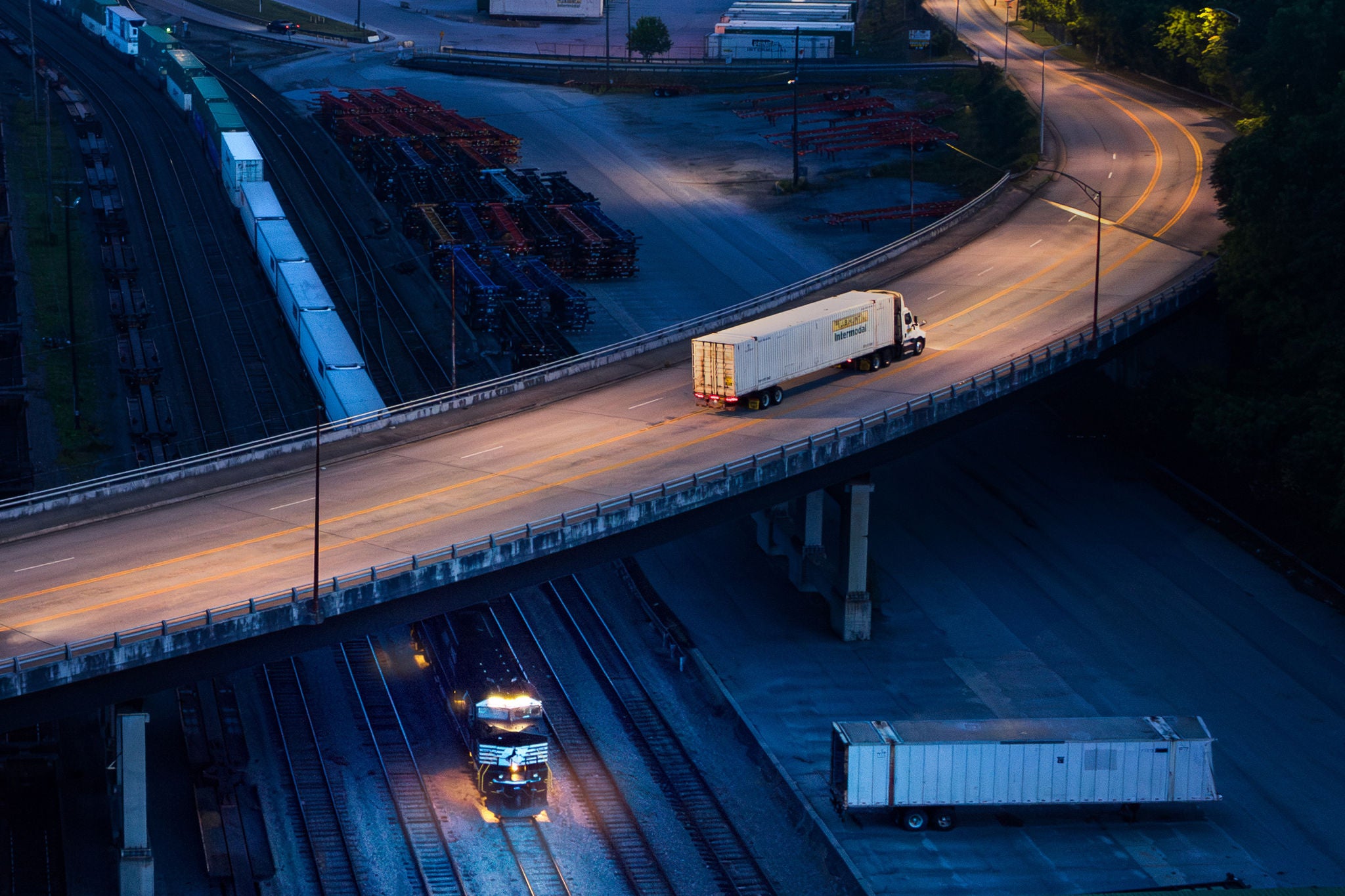 An intermodal container transport train moves along the tracks that stretch under a bridged freeway.